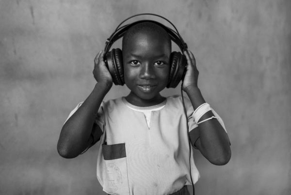 Portret afrykańskiego chłopca. Chłopiec jest uśmiechnięty na głowie ma założone słuchawki stereofoniczne, które po obu stronach dłońmi