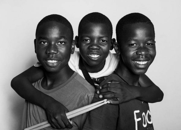 Portret trzech afrykańskich chłopców, Chłopcy są uśmiechnięci jeden z nich obejmuje dwóch pozostałych, a w ręce trzyma pałeczki perkusyjne.