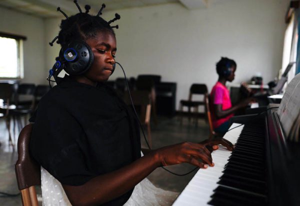 Zdjęcie przedstawia afrykańską dziewczynkę uderzającą w klawisze pianina, na uszach ma słuchawki stereofoniczne. W tle widać także drugą dziewczynkę grającą na drugim pianinie.