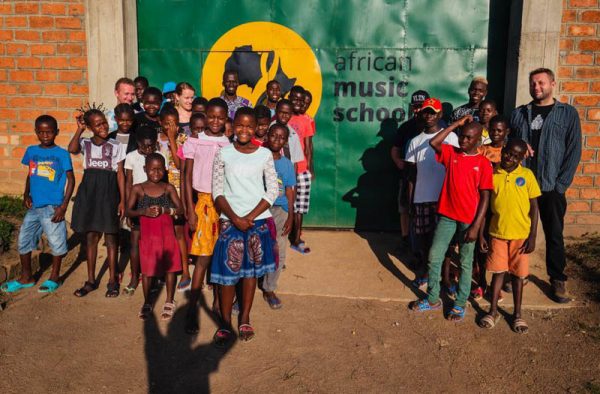Zdjęcie przedstawia grupę afrykańskich dzieci na tle ceglanej ściany, na której umieszczony został duży baner z logotypem " African music school"