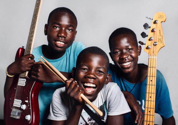 Zdjęcie przedstawia trzech uśmiechniętych afrykańskich chłopców. Jeden z nich trzyma gitarę basową, drugi pałeczki perkusyjne a trzeci gitarę elektryczną.