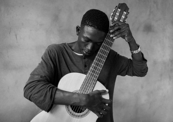 Czarno biały portret afrykańskiego chłopca, który jedną ręką obejmuje pudło rezonansowe gitary klasycznej, drugą ręką zaś trzyma jej gryf, do którego przytula się też głową.