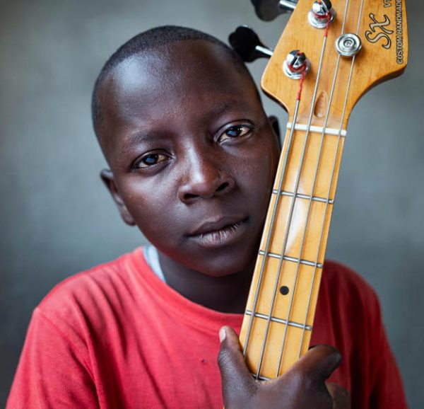 Portret afrykańskiego chłopca, który trzyma w jednej ręce blisko twarzy gryf gitary elektrycznej, jest do niego jakby przytulony.