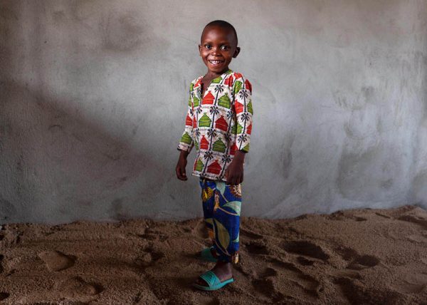 Zdjęcie przedstawia afrykańskiego chłopca w wieku około 4-5 lat, który uśmiecha się. Ma na sobie kolorowe materiałowe spodnie w słoneczniki i bluzkę w afrykańskie chatki i palmy, na jego nogach są klapki.