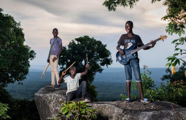 Zdjęcie przedstawia 3 afrykańskich chłopców na skale. Jeden z siedzi z uniesionymi rękami, w których trzyma pałeczki perkusyjne, a pozostali trzymają gitary elektryczne. Chłopcy mają otwarte usta jakby śpiewali lub krzyczeli.